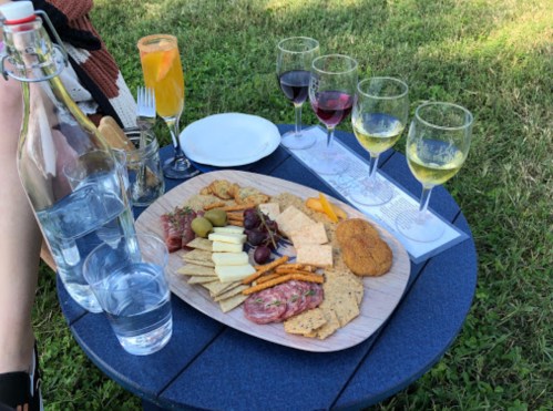 A wooden platter with assorted cheeses, meats, crackers, and olives, alongside glasses of wine and a colorful drink.