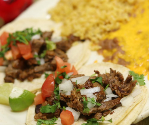 Two tacos filled with seasoned meat, topped with onions and tomatoes, served with rice and beans on the side.