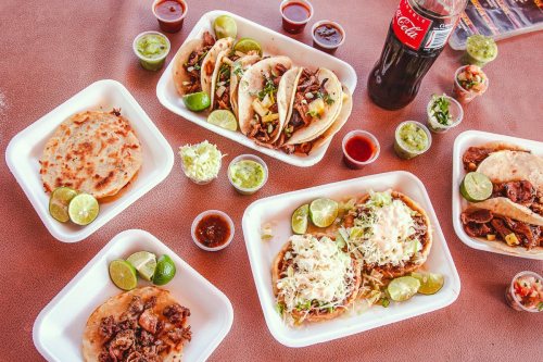 A spread of tacos and quesadillas with lime wedges, sauces, and a bottle of Coca-Cola on a table.