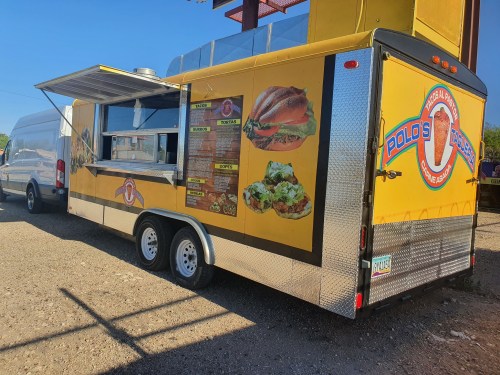 A bright yellow food truck with a menu displayed on the side, parked in a gravel area under a clear blue sky.