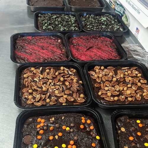 A variety of brownies in black trays, topped with nuts, candies, and colorful swirls, arranged on a table.