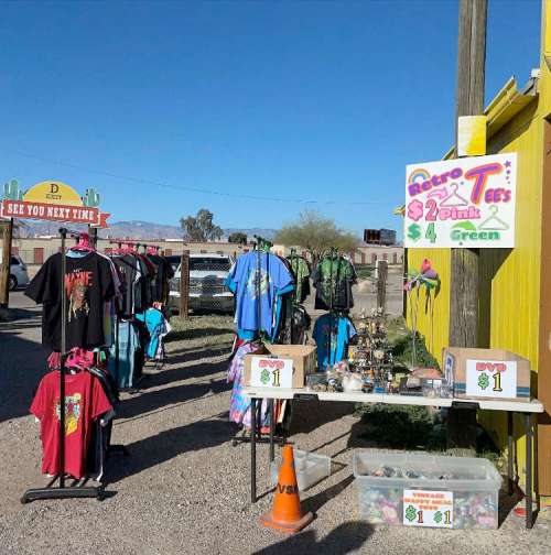 A colorful display of T-shirts for sale outside a shop, with a sign advertising prices and a table of items nearby.