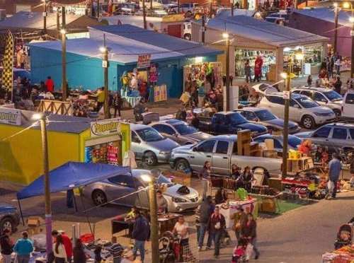 A bustling outdoor market with colorful stalls, parked cars, and people shopping in the evening.