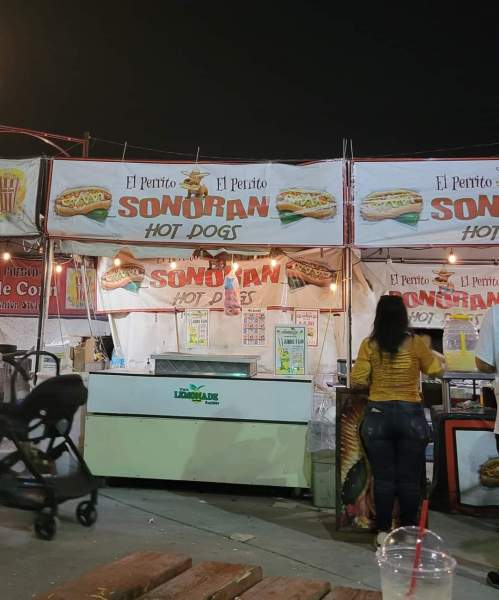 A food stall named "El Perrito Sonoran Hot Dogs" with colorful signage, serving hot dogs at night.