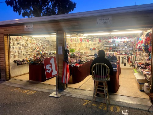 A person sits at a stall outside a shop filled with colorful items, with a sign displaying "$1" and an American flag.