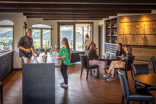 A modern winery interior with a bartender serving a customer, while others enjoy drinks at tables. Scenic views visible.