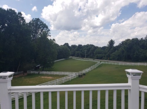 A scenic view of a green landscape with white fences and trees under a partly cloudy sky.