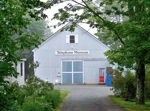 A blue barn-like building with a sign reading "Telephone Museum," surrounded by trees and a gravel path.