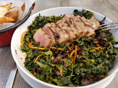 A bowl of mixed greens salad topped with sliced grilled tuna and carrots, alongside a basket of bread.