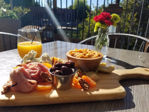 A wooden platter with assorted meats, cheese, nuts, and dried fruits, alongside a glass of orange juice and flowers.