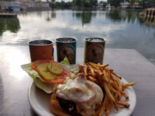 A plate with a burger topped with cheese and bacon, fries, and a salad, with drinks and a scenic lake in the background.