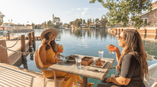 Two women enjoy drinks and food by a serene lakeside, with a sunny sky and charming homes in the background.