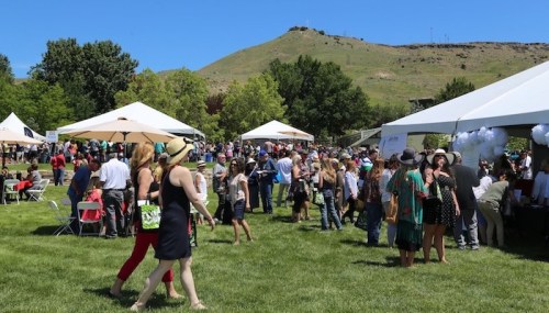 A lively outdoor event with crowds, tents, and a scenic hillside in the background on a sunny day.