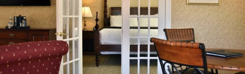 A hotel room with a bed visible through glass-paneled doors, featuring a desk and chair in the foreground.