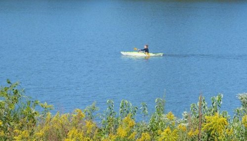 A person paddles a yellow kayak on a calm blue lake, surrounded by green and yellow foliage.