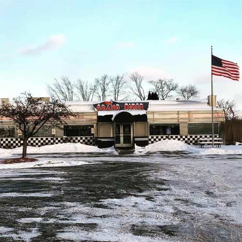 A classic diner with a sign reading "Grand Diner," surrounded by snow and an American flag waving in the background.