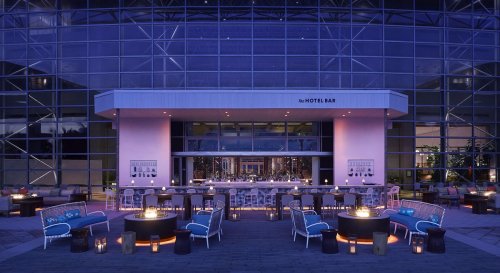 Outdoor hotel bar at dusk, featuring stylish seating and glowing fire pits, surrounded by modern architecture.