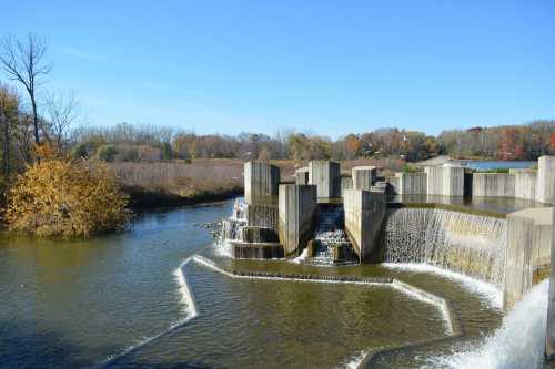 A concrete waterfall structure flows into a calm river, surrounded by trees and autumn foliage under a clear blue sky.