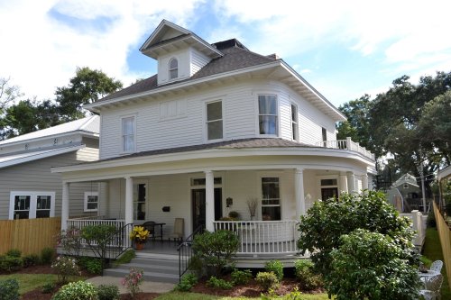 A charming two-story white house with a wraparound porch, surrounded by greenery and a clear blue sky.