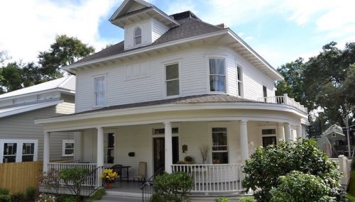 A charming two-story white house with a wraparound porch and a turret, surrounded by greenery.