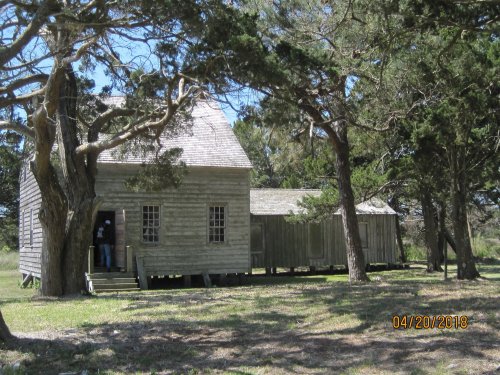 A weathered wooden house surrounded by trees, with a smaller building nearby, set in a grassy area.