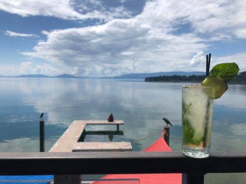 A refreshing cocktail sits on a railing, overlooking a calm lake and blue sky with fluffy clouds.