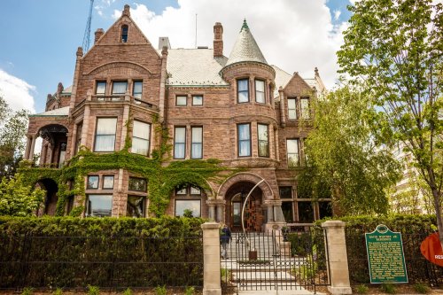 A large, historic stone mansion with ivy, featuring a turret and intricate architectural details, surrounded by greenery.