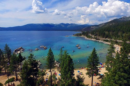 A scenic view of a beach by a lake, surrounded by trees and mountains, with people enjoying the water and sun.
