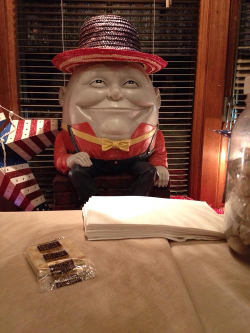 A large, smiling statue of a man in a hat and bow tie sits on a table with napkins and snacks nearby.