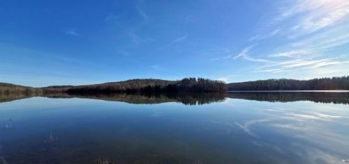 A serene lake reflecting a clear blue sky and surrounding trees, with gentle hills in the background.
