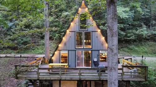 A cozy A-frame cabin nestled in the woods, featuring large windows and a spacious deck surrounded by trees.
