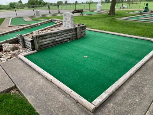 A mini-golf course with green turf, wooden borders, and a stone marker in the background.