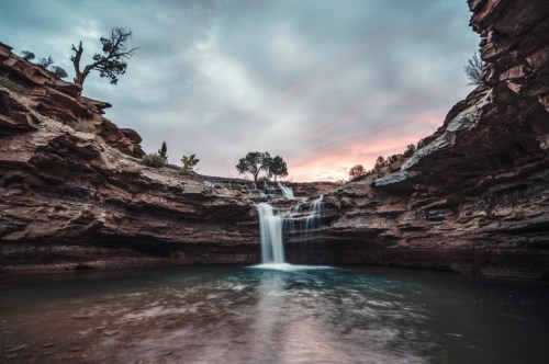 A serene waterfall cascades into a calm pool, surrounded by rocky cliffs and a cloudy sky at sunset.