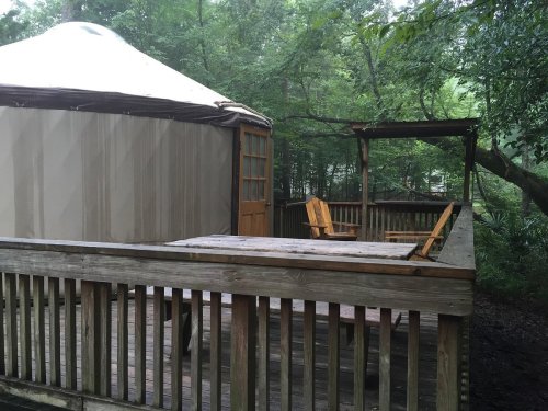 A wooden deck with two chairs beside a yurt, surrounded by lush green trees in a tranquil setting.