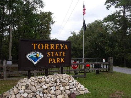 Sign for Torreya State Park with a flagpole and trees in the background, featuring a stone pile in the foreground.