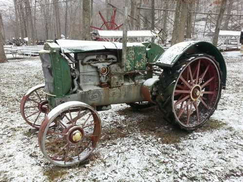 An old, green tractor with red wheels sits in a snowy forest clearing.