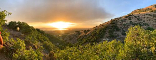 A panoramic view of a sunset over a valley, framed by green hills and trees.