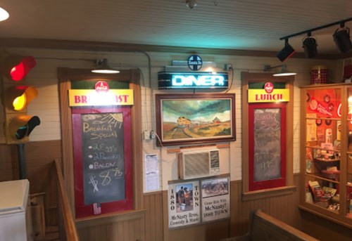 Interior of a diner featuring breakfast and lunch menus on chalkboards, with a neon "Diner" sign and colorful decor.