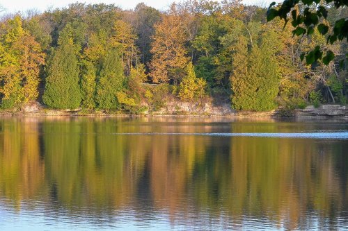 A serene lake reflecting colorful autumn trees along its shore, with calm water and a clear sky.