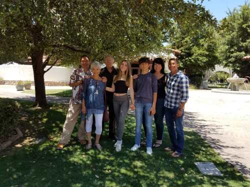 A group of seven people stands together outdoors under a tree, smiling in a sunny garden setting.