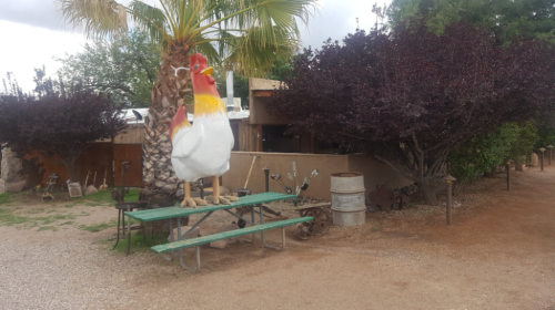 A large chicken statue stands on a picnic table surrounded by trees and rustic decor in a garden setting.