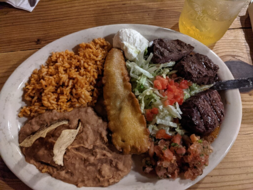 A plate of Mexican food featuring rice, refried beans, grilled meat, a fried item, lettuce, tomatoes, and sour cream.