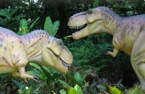 Two lifelike dinosaur sculptures, resembling T. rex, face each other amidst lush green foliage.