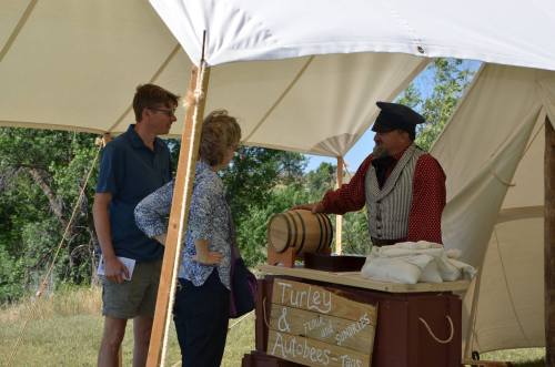 A vendor in period attire talks to two visitors at a tented event, with a barrel and sign for "Turley & Autobrews."