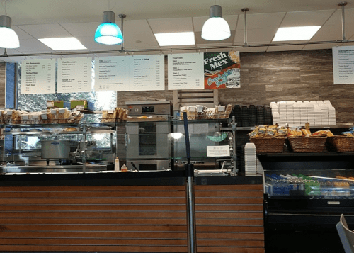 A café counter with a menu board, food displays, and shelves of cups and snacks in a bright, modern setting.