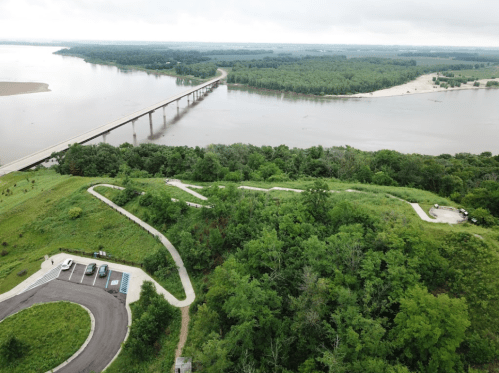 Aerial view of a river with a bridge, surrounded by lush greenery and a winding path near a parking area.