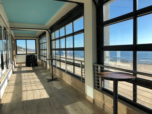 Bright, modern interior with large windows overlooking a beach and ocean, featuring wooden floors and small tables.
