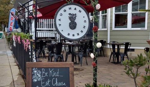 A cozy outdoor dining area with tables, a sign for Vaughan Cheese, and a chalkboard with the message "Be Kind... Eat Cheese."