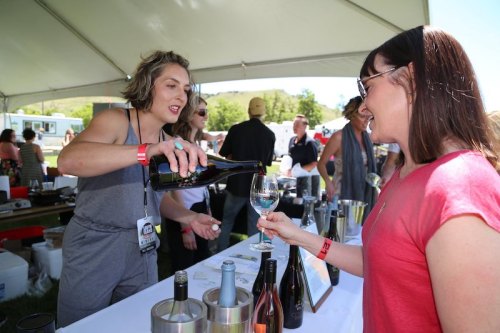 A woman pours wine into a glass for a guest at an outdoor event under a tent.