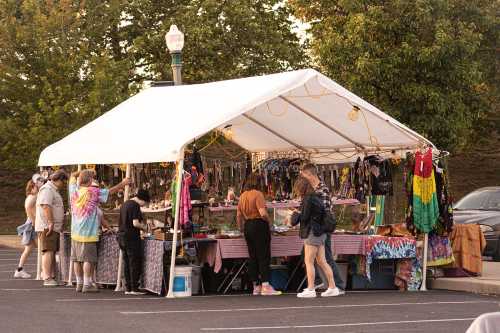 A colorful market tent with people browsing handmade crafts and jewelry in a parking lot during sunset.